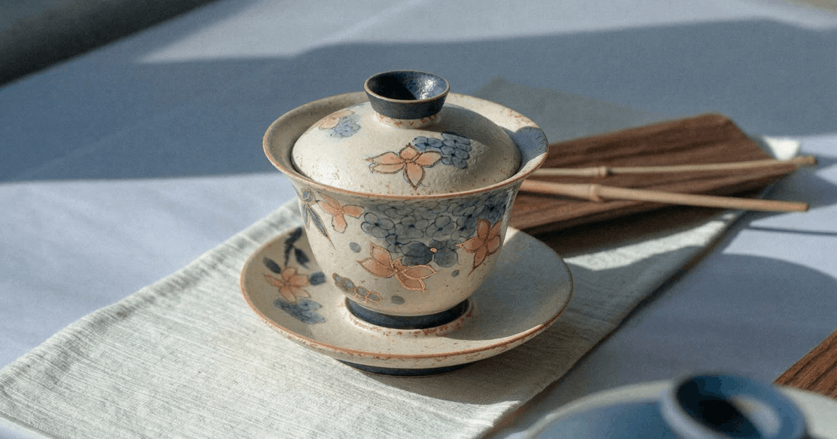 Chinese ceramic gaiwan tea set with hand-painted floral pattern, placed on linen cloth in natural sunlight, traditional gongfu tea brewing scene