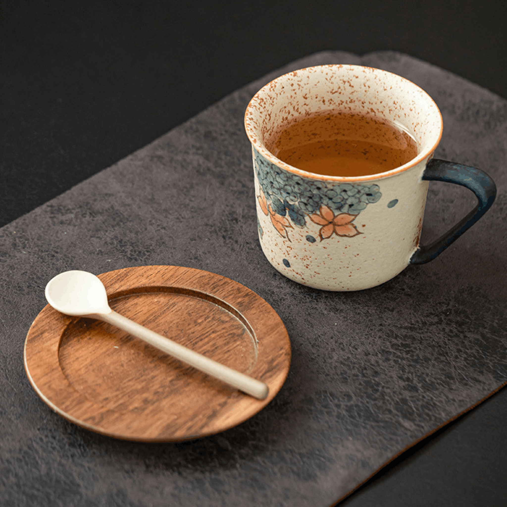 Ceramic mug with floral design on a wooden coaster with a spoon, all on a dark surface.