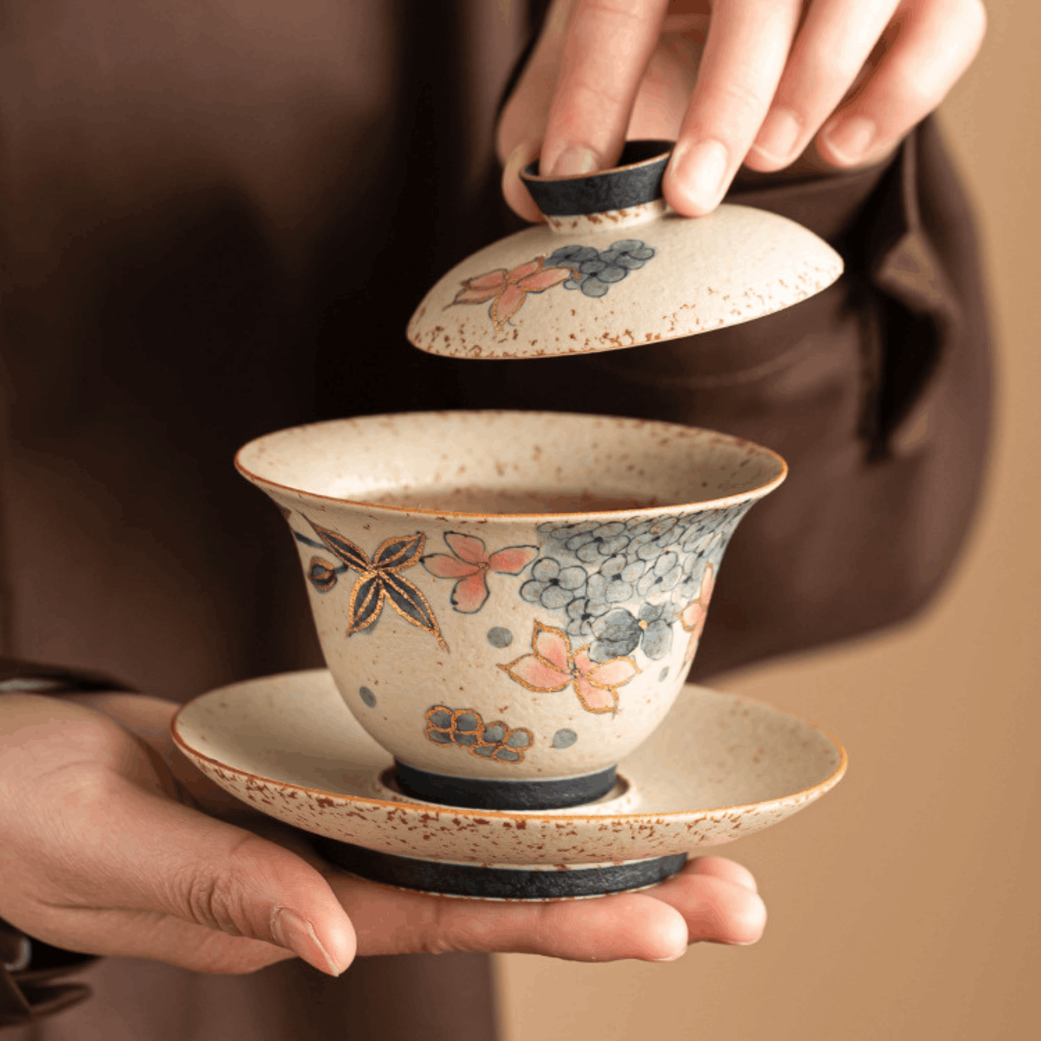 Ceramic gaiwan with floral design held by a person against a brown background