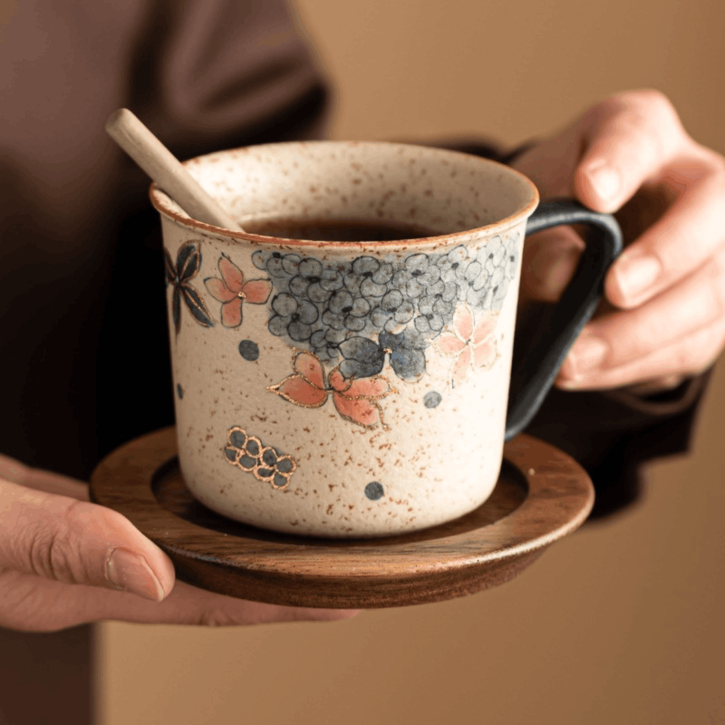 Ceramic mug with floral design on a wooden saucer held by hands against a brown background