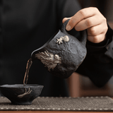 Person pouring tea from a black ceramic teapot into a matching bowl on a dark surface.